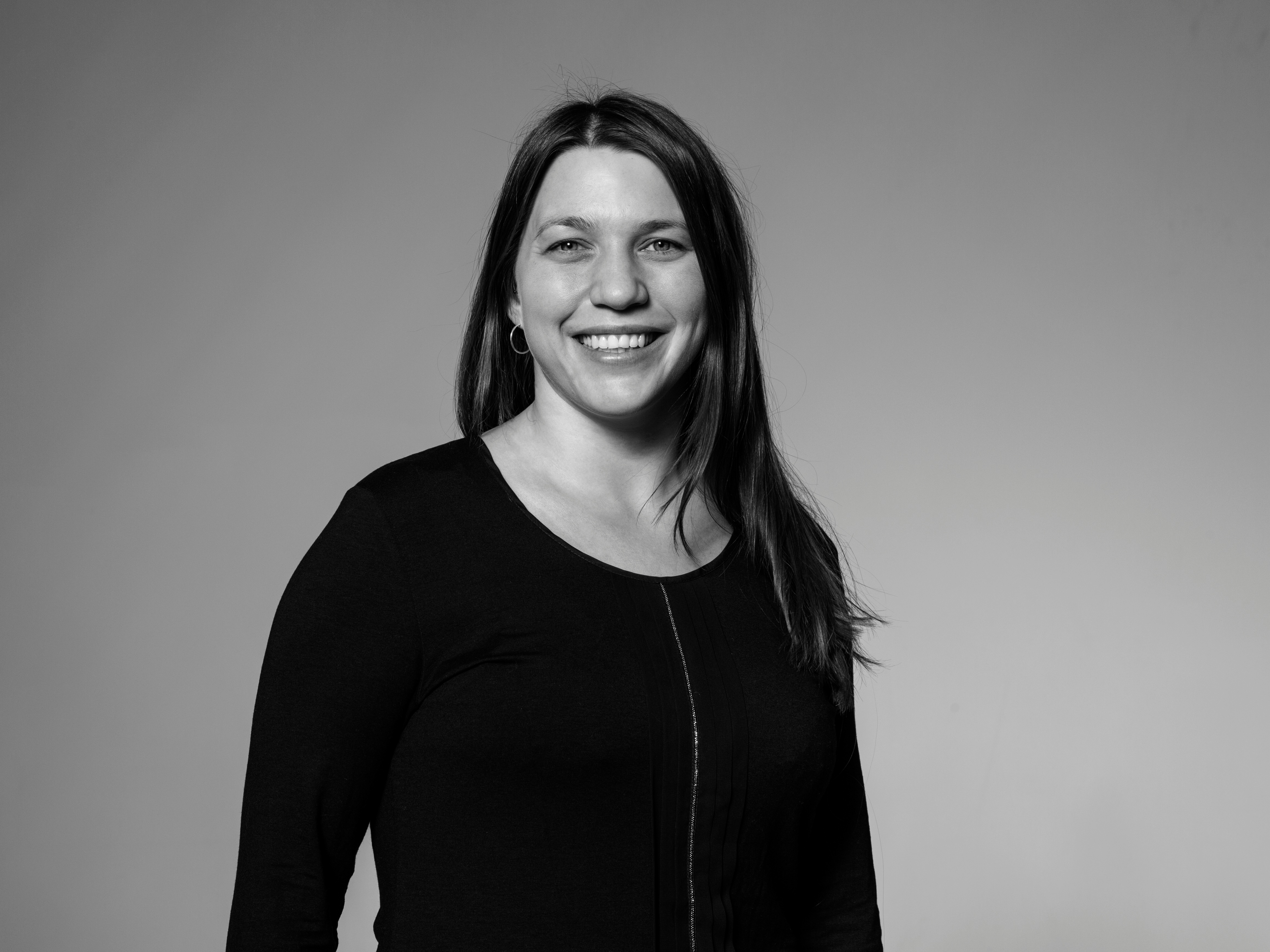 Sort-hvitt portrett av en smilende kvinne med langt, rett hår. Hun ser rett inn i kameraet med et varmt og imøtekommende smil. Kvinnen er iført en enkel sort genser, og bildet er i en sekskantet ramme, noe som tilfører et stilfullt og moderne element til presentasjonen.