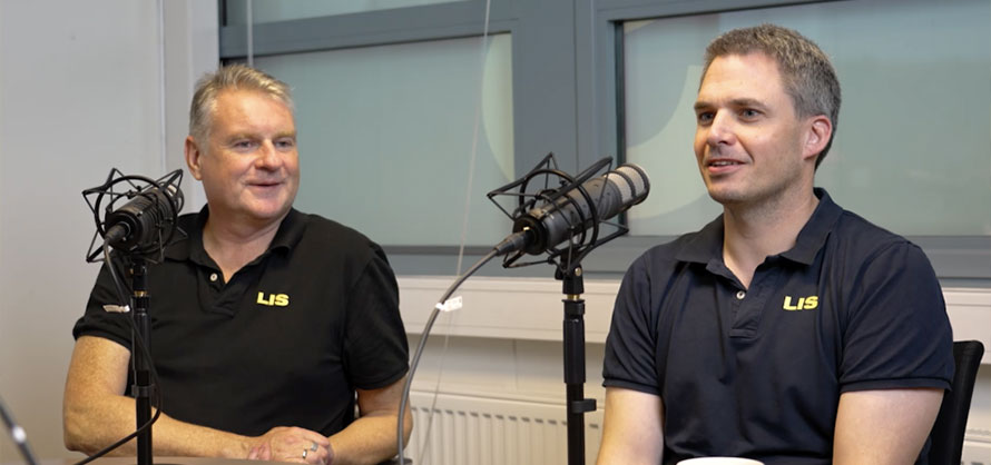 To menn i svarte poloskjorter med logoen 'LIS' sitter i et radiostudio og snakker i mikrofoner. Den ene mannen smiler mens den andre ser på ham og lytter oppmerksomt. Bakgrunnen viser et nøytralt og teknisk studioinnredning