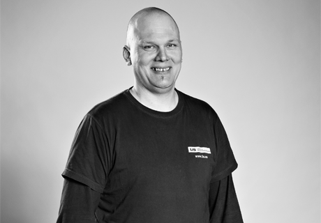 Et portrett av en mann i et sekskantet format. Han smiler og har på seg en sort t-skjorte med et navneskilt merket