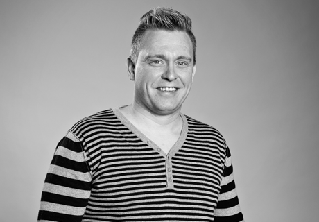 Et portrett av en mann med et vennlig smil, kledd i en stripete genser med en V-hals. Bildet er i svart-hvitt, i et sekskantet format, som gir et moderne og stilrent uttrykk.