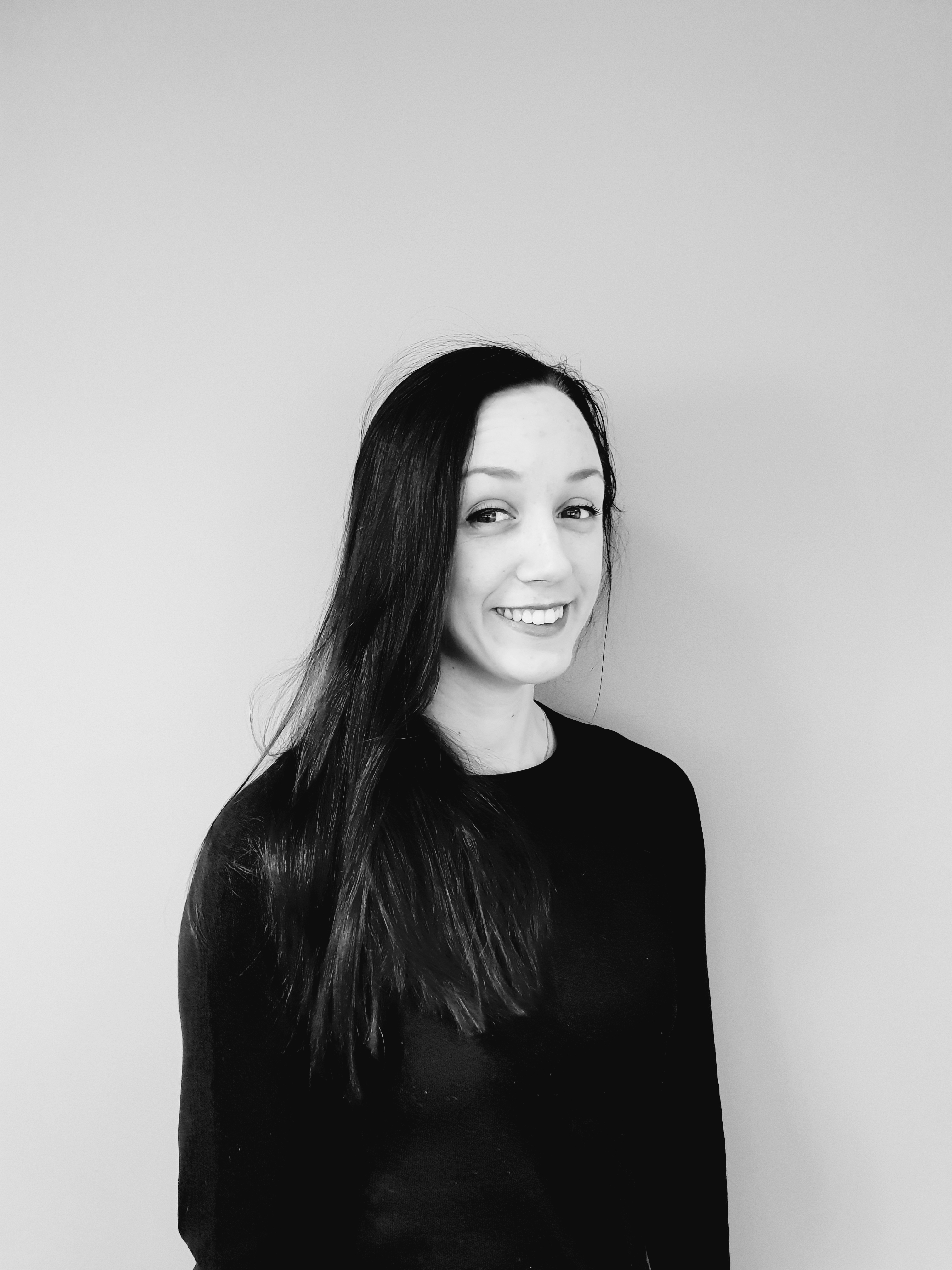 Et sort-hvitt portrett av en smilende ung kvinne. Hun ser direkte inn i kameraet med et vennlig og åpent ansikt. Hennes lange, mørke hår er slakt og faller naturlig. Hun har på seg en enkel, sort topp, og bakgrunnen er nøytral og gir ingen distraksjoner, som setter hele fokuset på hennes uttrykksfulle ansikt